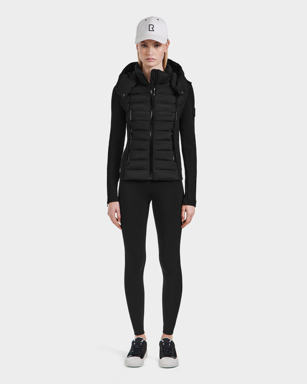 A person stands facing forward against a plain white background, wearing a Rudsak white baseball cap, a black lightweight short jacket over a fitted black long-sleeve top, black leggings, and Rudsak MABS - 8124825 Black sneakers with white soles.