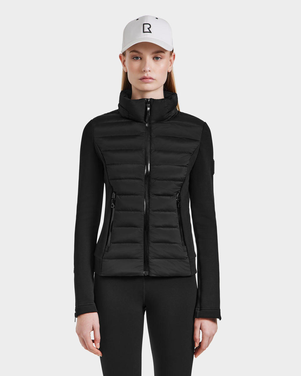 A woman wears the Rudsak MABS - 8124825 Black lightweight short jacket, black pants, and a white baseball cap with a black R logo, standing facing forward against a plain light background.