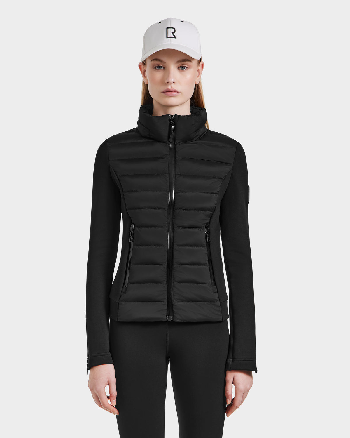 A woman wears the Rudsak MABS - 8124825 Black lightweight short jacket, black pants, and a white baseball cap with a black R logo, standing facing forward against a plain light background.