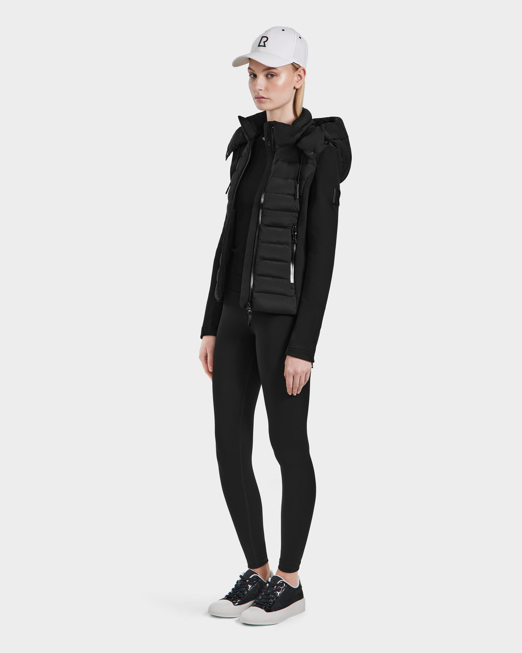 A person wearing a white baseball cap, a Rudsak MABS - 8124825 Black hooded puffer vest, black long-sleeve top, black leggings, and black sneakers stands against a plain white background.