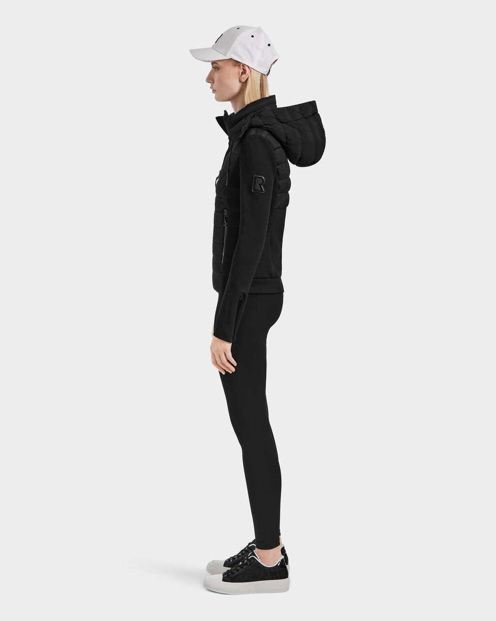 A person in a black Rudsak MABS - 8124825 Black womens jacket, black leggings, black sneakers, and a white baseball cap stands in profile against a plain white background.