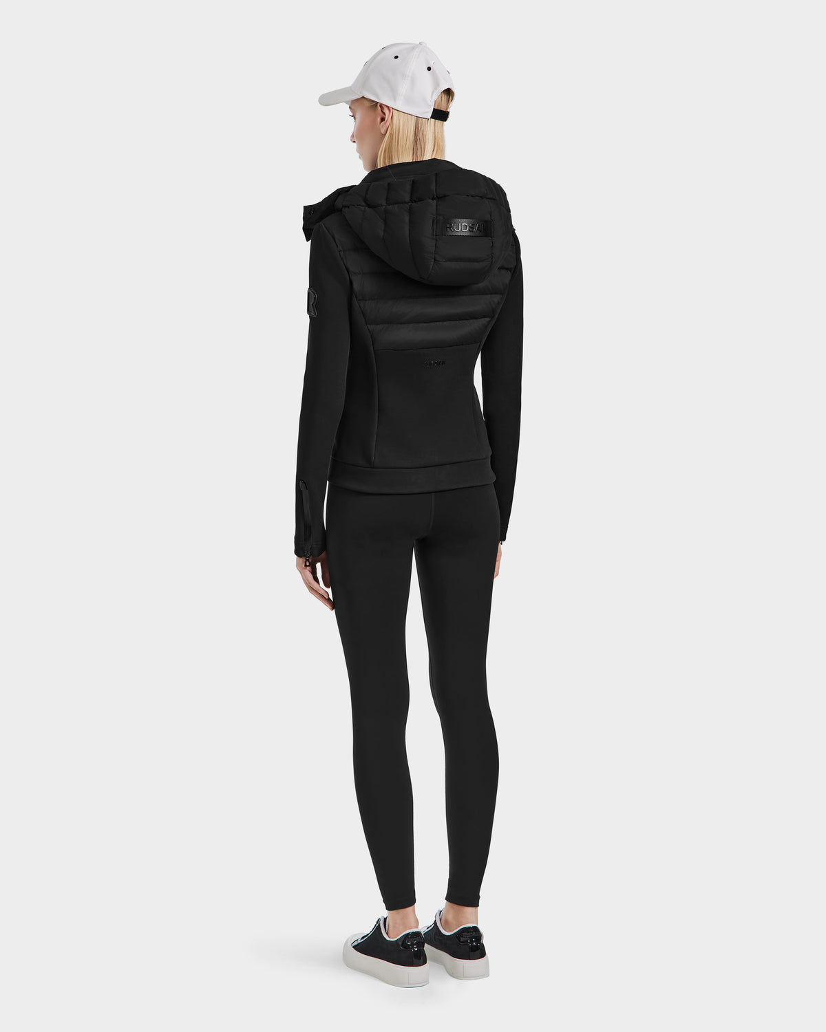 A person wearing the Rudsak MABS - 8124825 Black lightweight short jacket, black leggings, black and white sneakers, and a white baseball cap stands facing away against a plain white background.