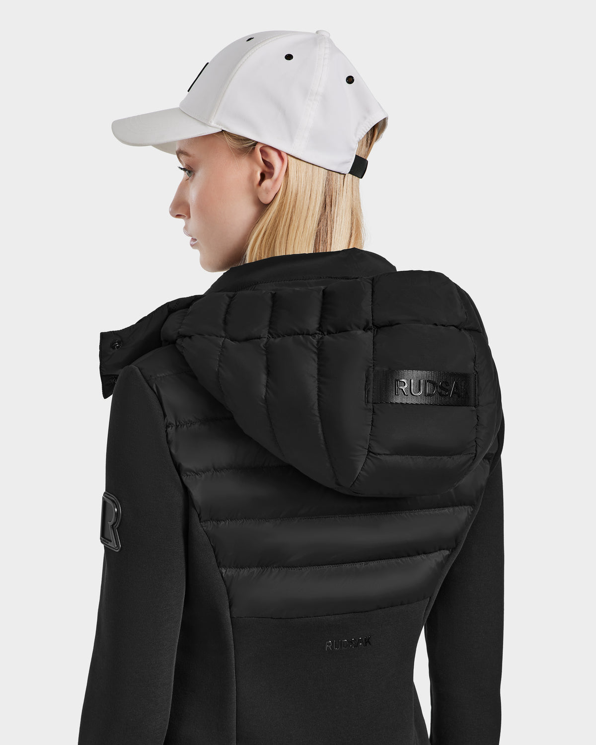 A blonde woman wears a white baseball cap and the Rudsak MABS - 8124825 Black womens lightweight short jacket with padded hood, shown from back and side against a plain background. Branding appears on the hood and sleeve.