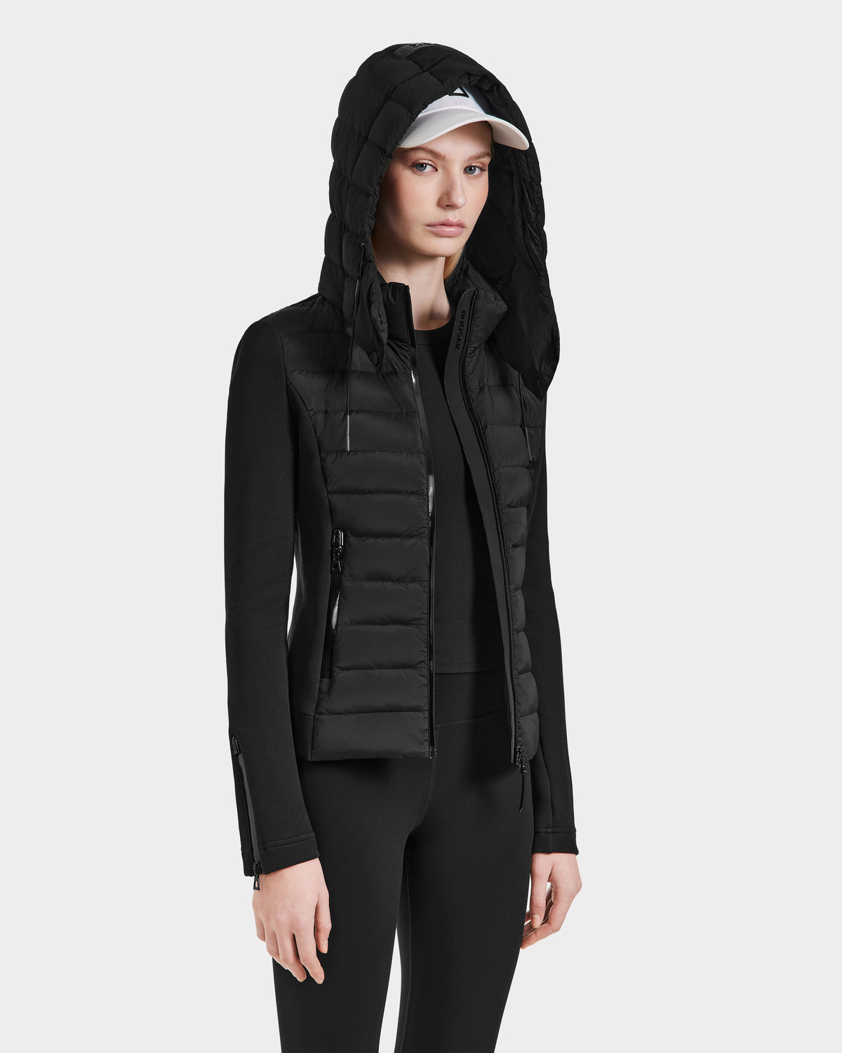 A woman wears the Rudsak MABS - 8124825 Black fitted women’s jacket with a hood, styled over a white baseball cap, black top, and black pants, standing against a plain white background.