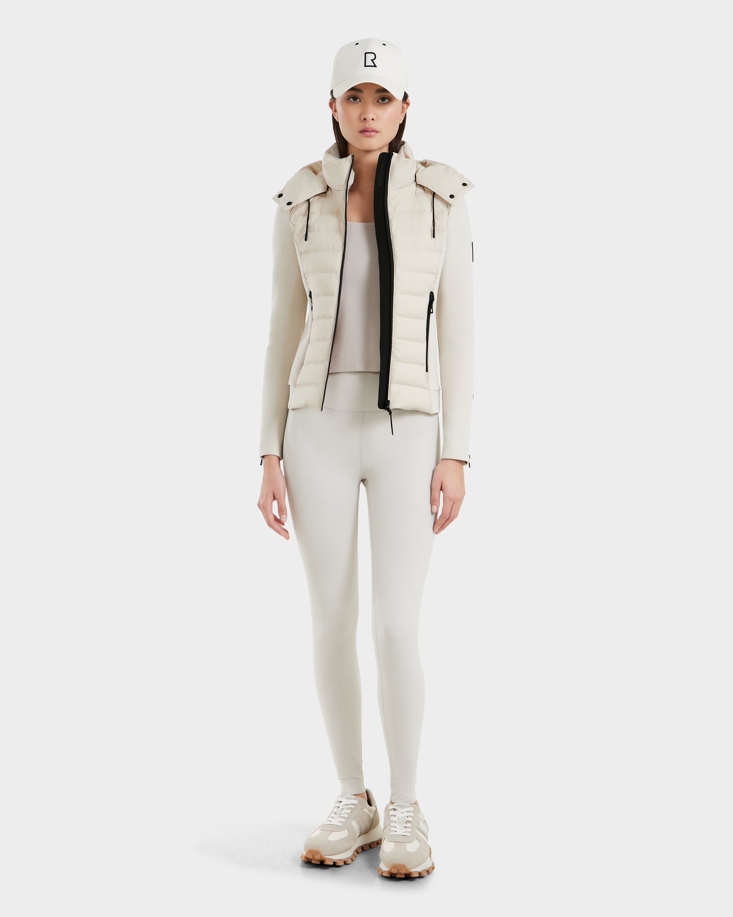 A woman faces forward against a plain white background, modeling the Rudsak MABS - 8125055 Vanilla women’s puffer jacket with RDS-certified down, paired with a white cap, light beige zip-up top, matching leggings, and beige sneakers.