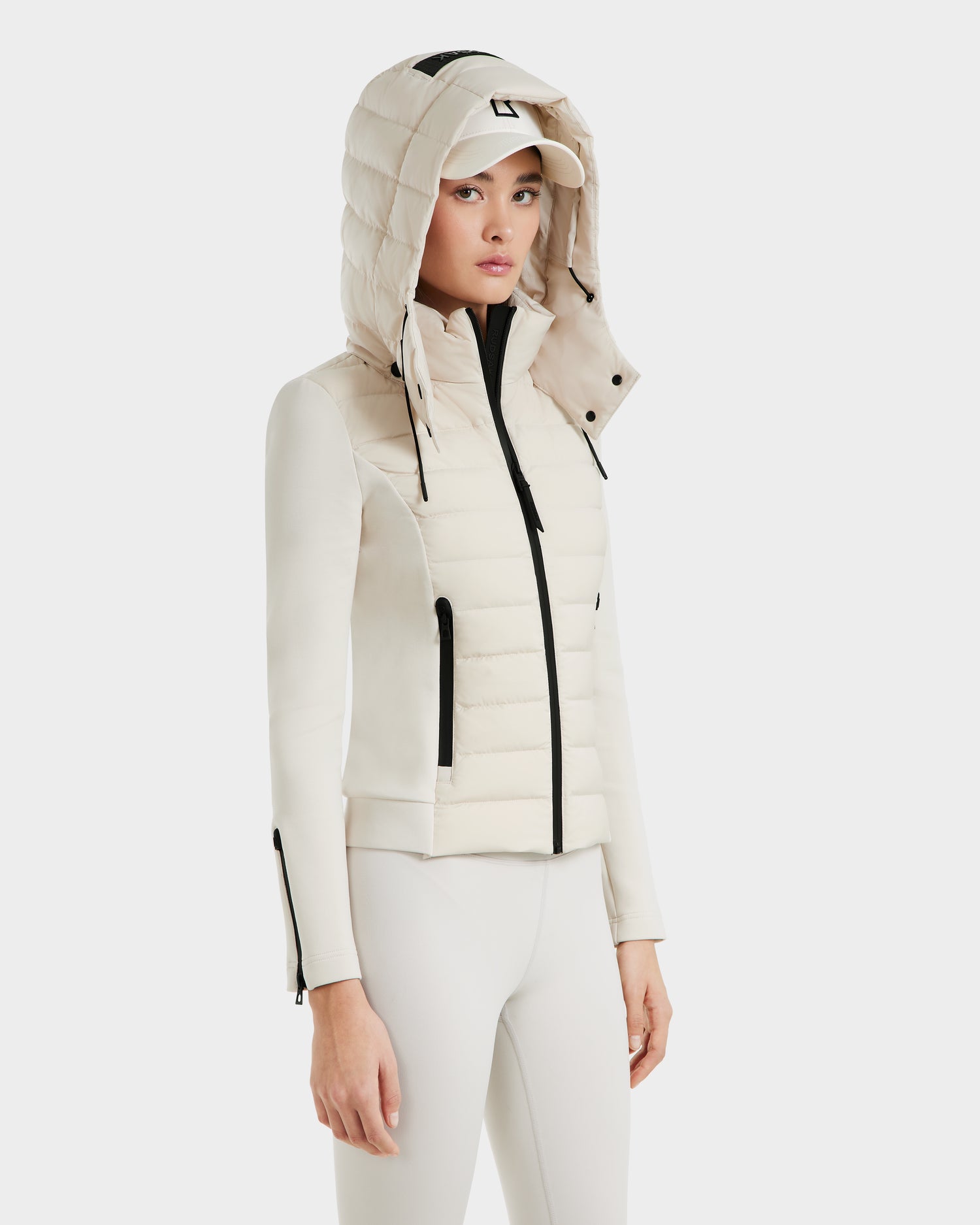 A woman wears the Rudsak MABS - 8125055 Vanilla, a fitted cream activewear jacket with quilted panels, black zippers, and RDS-certified down. She stands against a white background with the hood up over her matching cap and leggings.