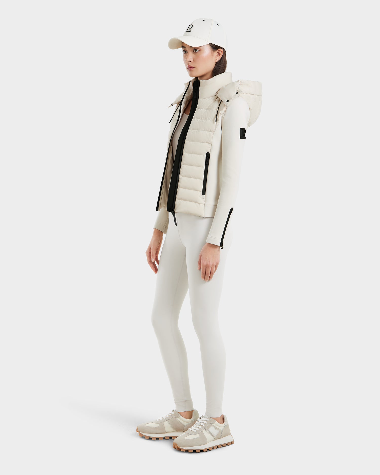 A woman wears Rudsak’s MABS - 8125055 Vanilla: an all-white athleisure look with a lightweight activewear jacket styled as a puffer, white cap, fitted long-sleeve top, leggings, and beige-white sneakers against a light background.