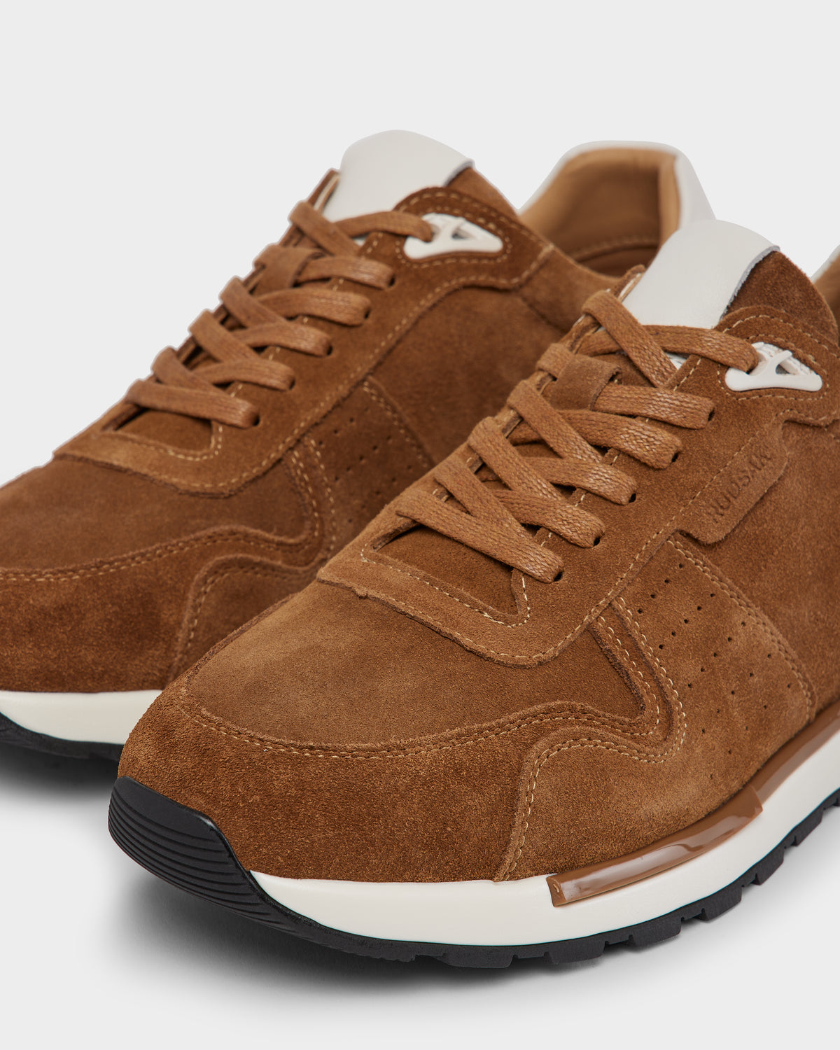 A close-up of the Rudsak MALONE S - 6225002 Almond men’s suede sneaker in brown, featuring tan laces, white accents on the tongue and sole, black rubber outsoles, and a modern design against a neutral background.