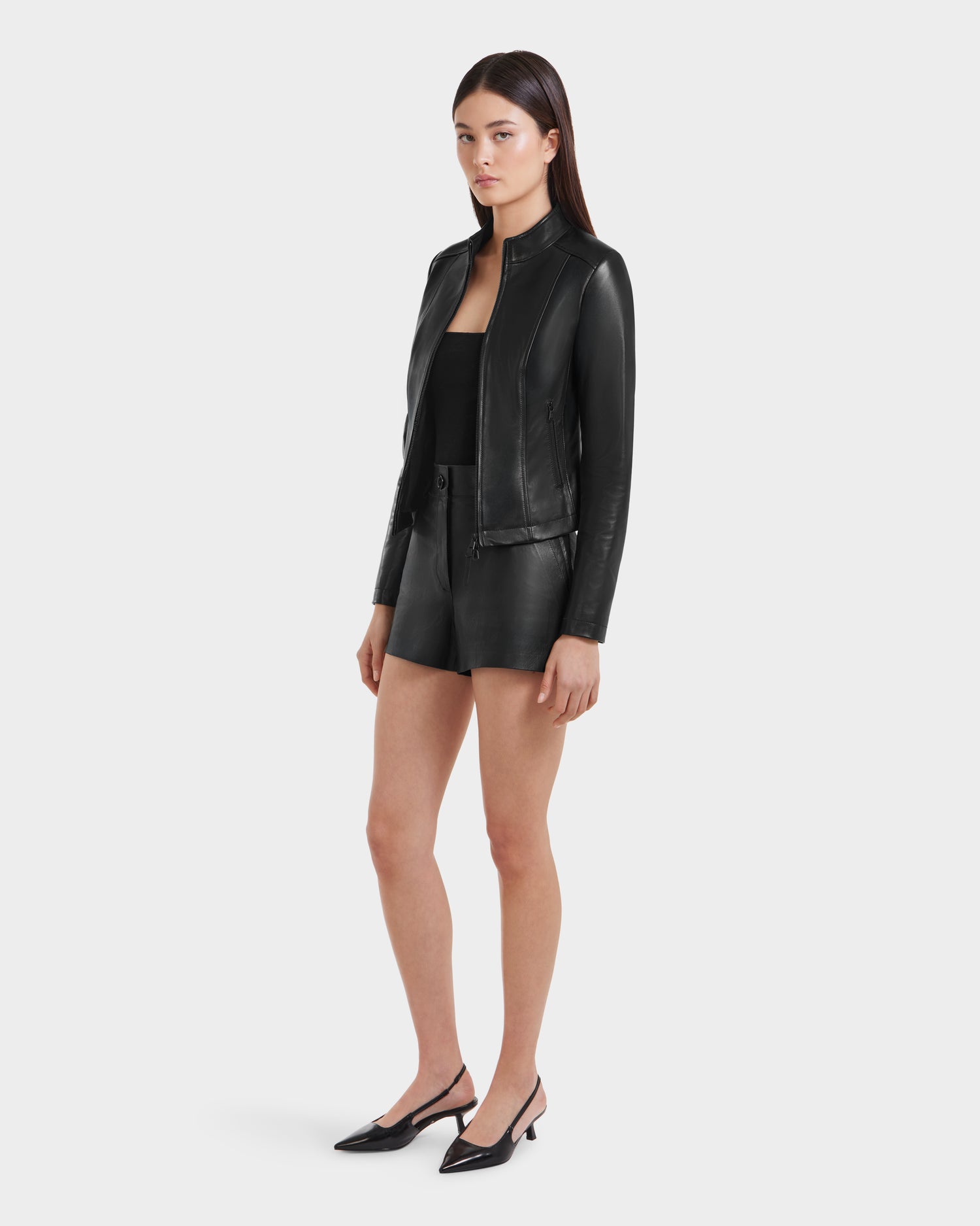 A woman stands against a plain background wearing the Rudsak MARIAH - L124511 Black moto biker jacket, black leather shorts, a black top, and low-heeled shoes. She looks slightly to the side with her arms relaxed at her sides.