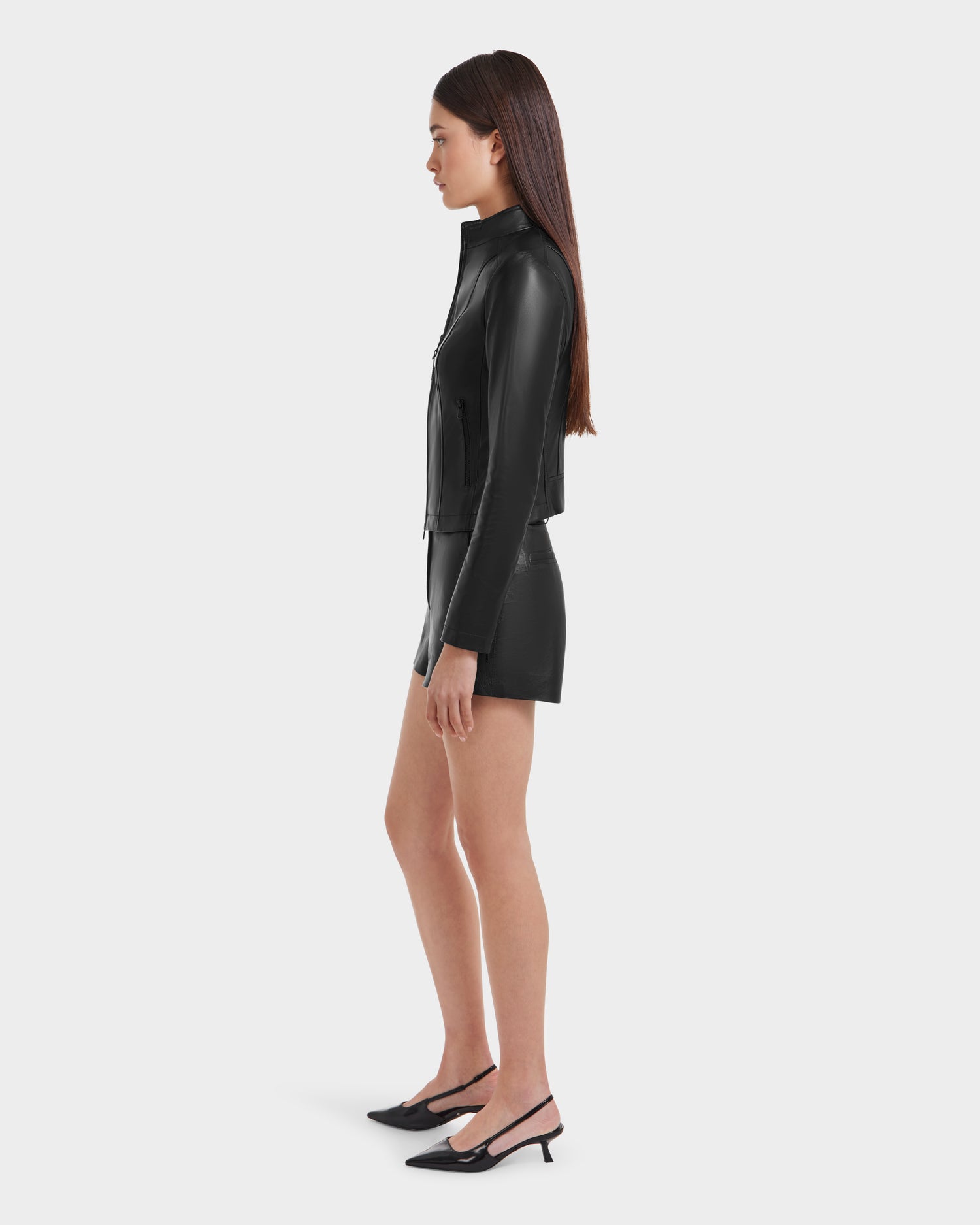 A woman in profile models the Rudsak MARIAH - L124511 Black women’s fitted leather jacket, a matching black leather mini skirt, and black slingback heels against a plain white background.