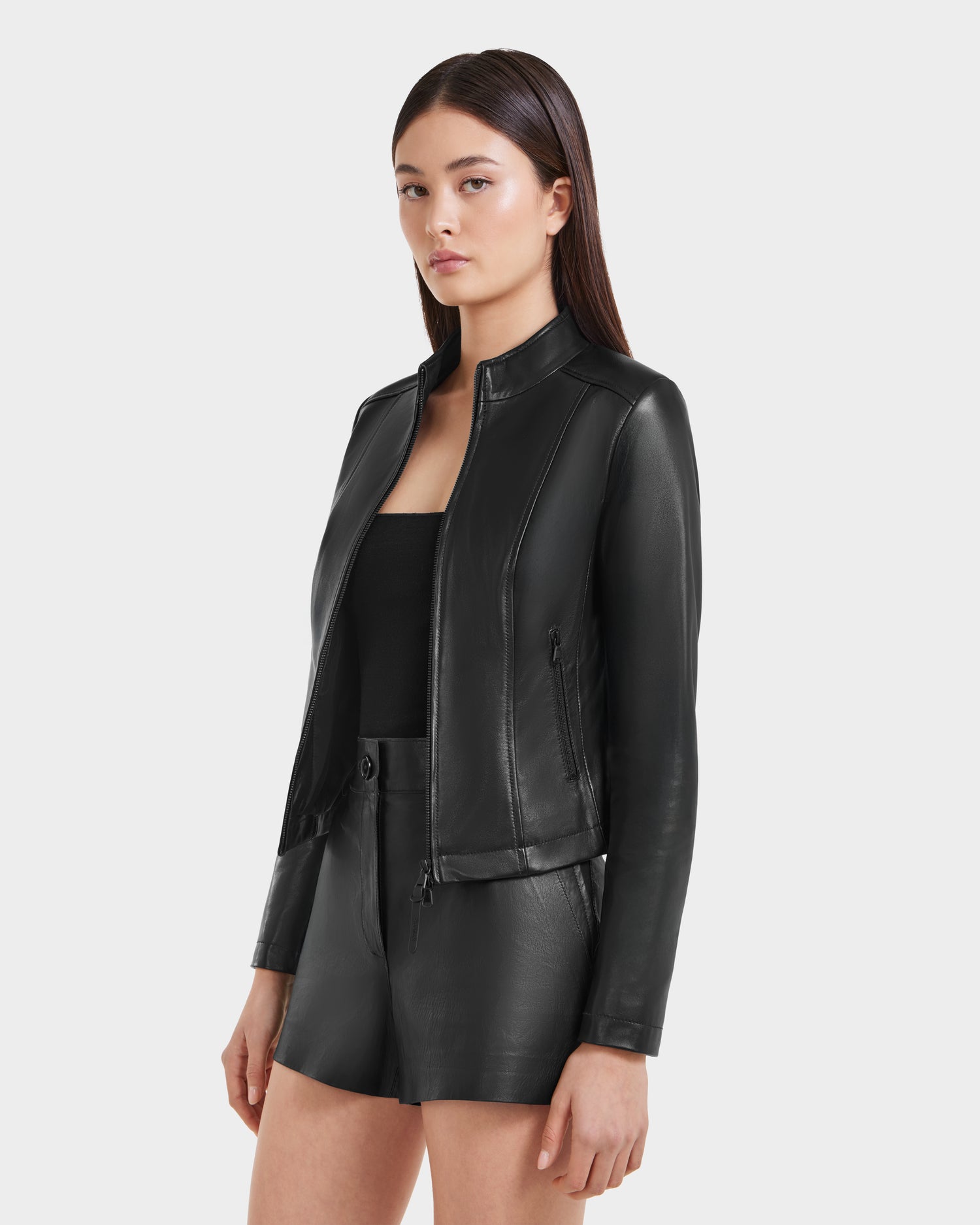 A woman with straight dark hair models the Rudsak MARIAH - L124511 Black fitted leather jacket over a black top and matching shorts, standing against a plain light background.