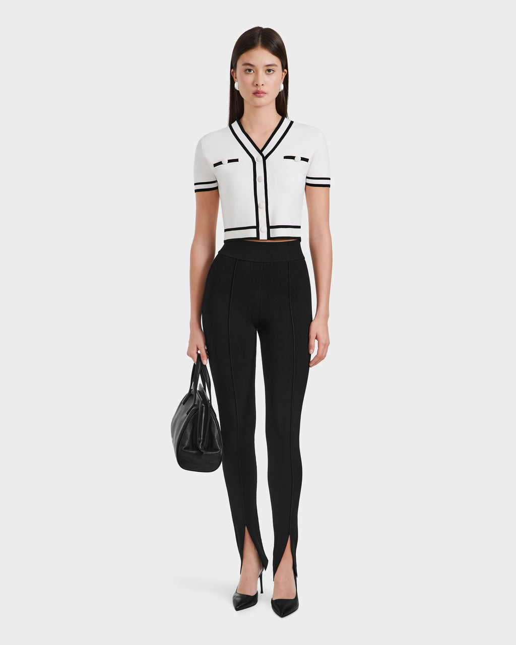 A woman faces forward, wearing a white short-sleeve cropped top with black trim, Rudsak MATHILDA - 8524515 Black ankle slit leggings, black heels, and holding a black handbag.