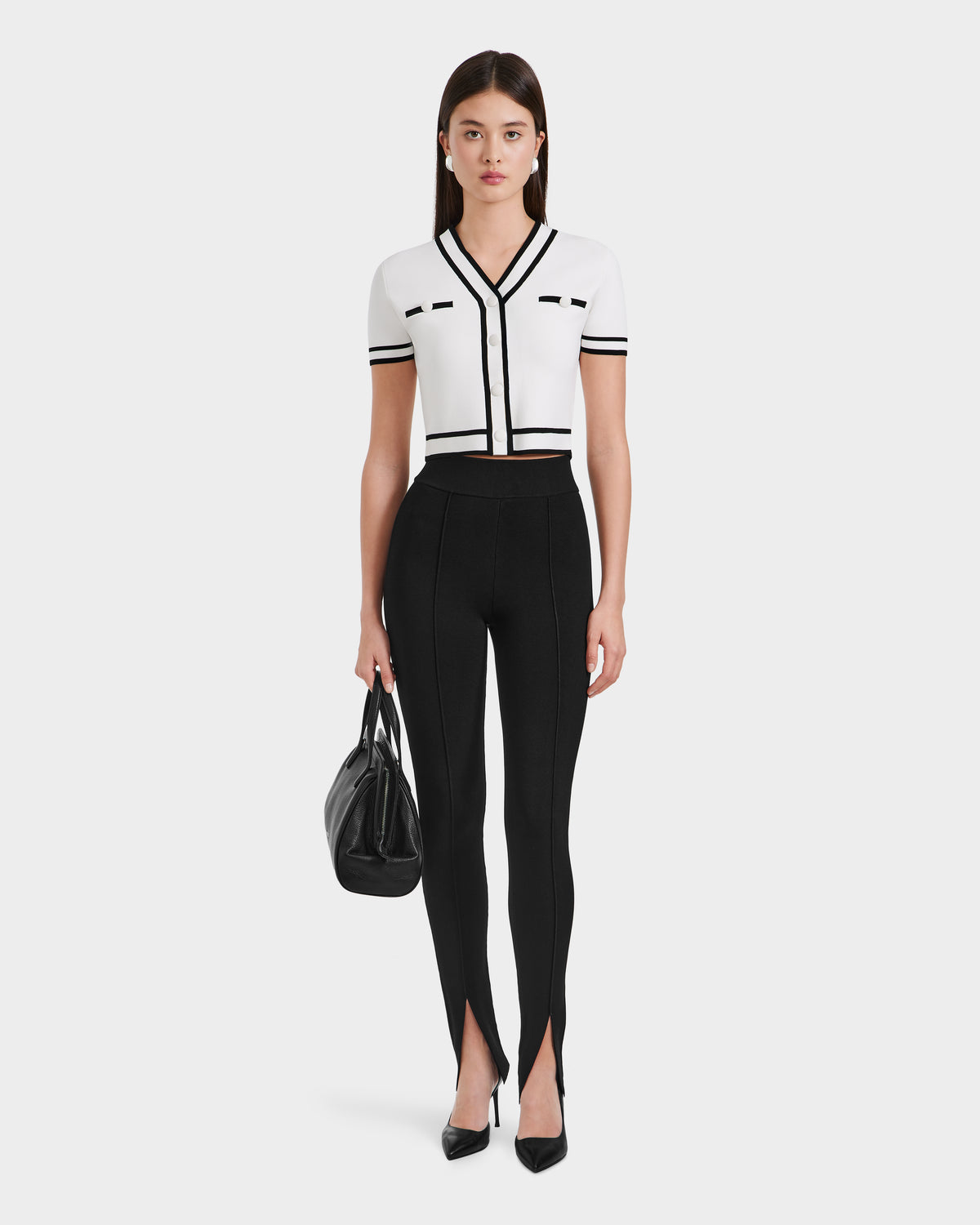 A woman faces forward, wearing a white short-sleeve cropped top with black trim, Rudsak MATHILDA - 8524515 Black ankle slit leggings, black heels, and holding a black handbag.