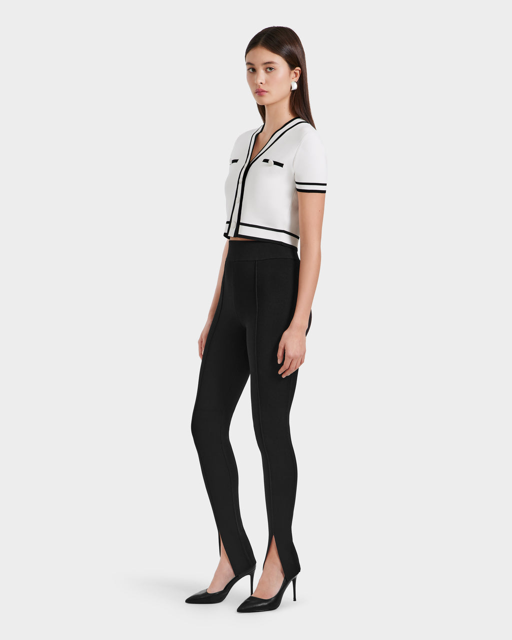 A woman stands in a studio wearing a white, short-sleeve, cropped cardigan with black trim and Rudsaks MATHILDA - 8524515 Black ankle slit leggings, paired with black heels. She faces slightly to the side.