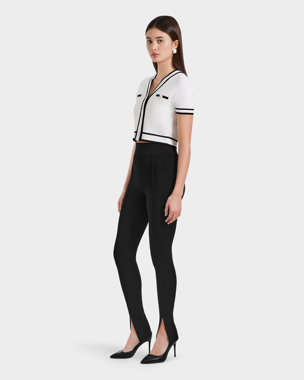 A woman stands in a studio wearing a white, short-sleeve, cropped cardigan with black trim and Rudsaks MATHILDA - 8524515 Black ankle slit leggings, paired with black heels. She faces slightly to the side.
