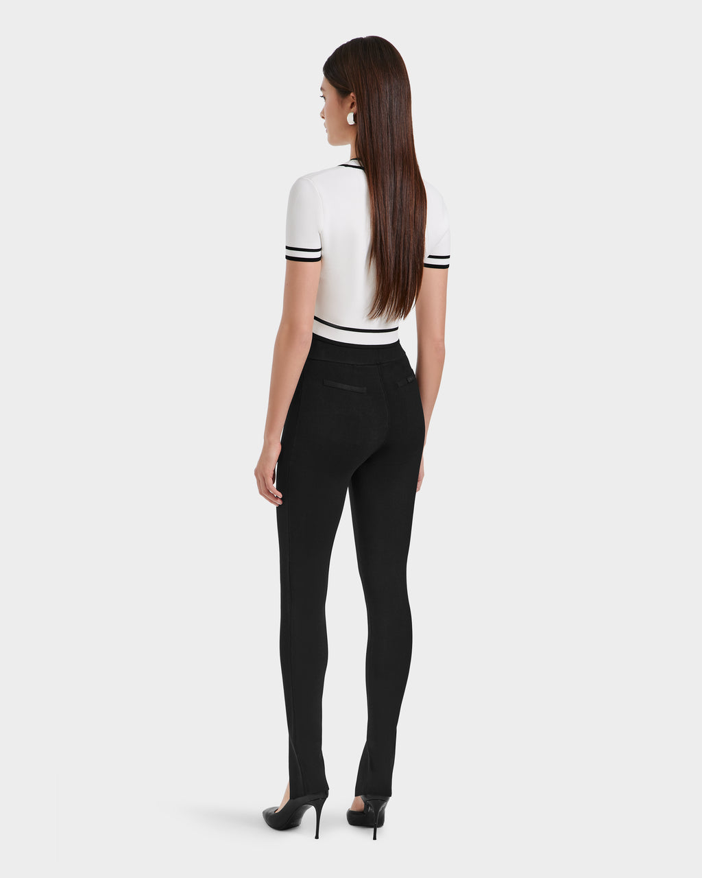 A woman faces away in a short-sleeve white top with black striped details, paired with Rudsaks MATHILDA - 8524515 Black womens knit leggings and black heels, standing against a plain light background.