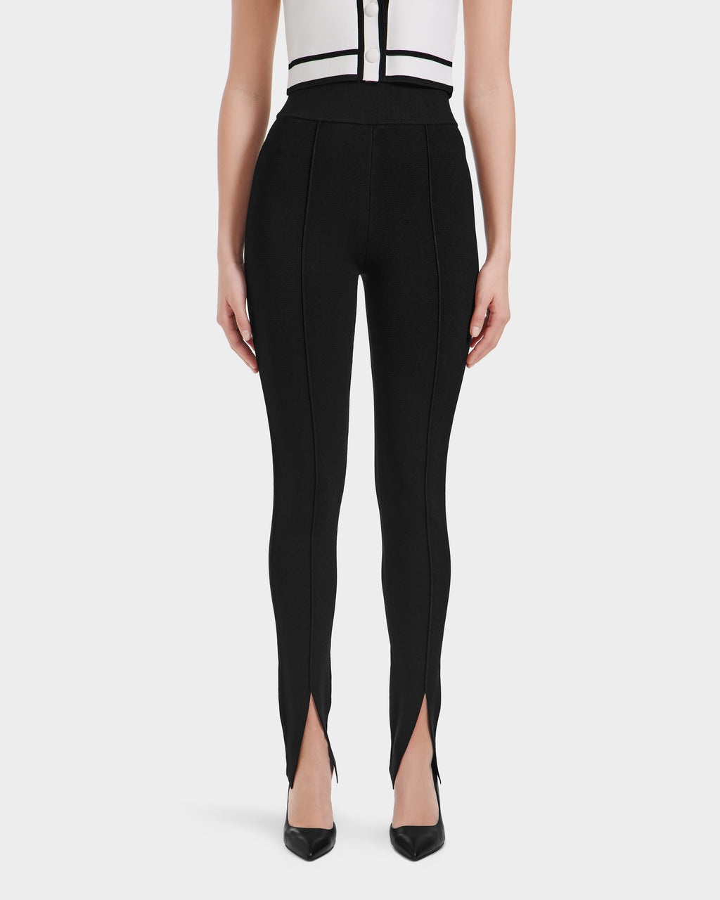A woman stands facing forward, wearing Rudsak’s MATHILDA - 8524515 Black high-waisted leggings with front ankle slits, paired with black pointed heels and a sleeveless white top featuring black accents.