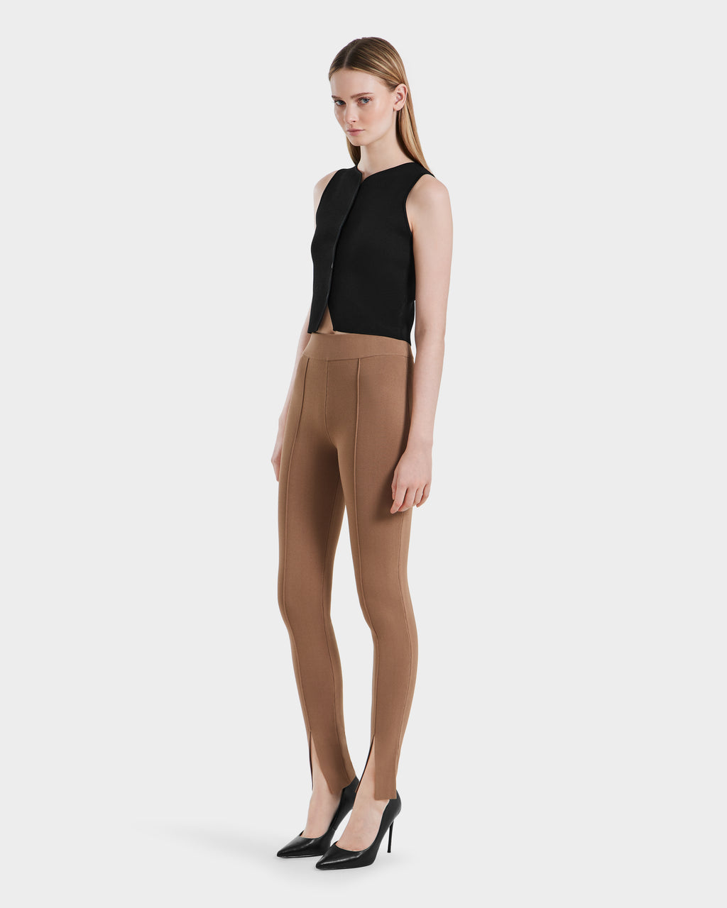 A woman stands against a plain background wearing a sleeveless black top, Rudsak MATHILDA - 8524515 Camel high-waisted brown ankle slit leggings, and black high-heeled shoes.