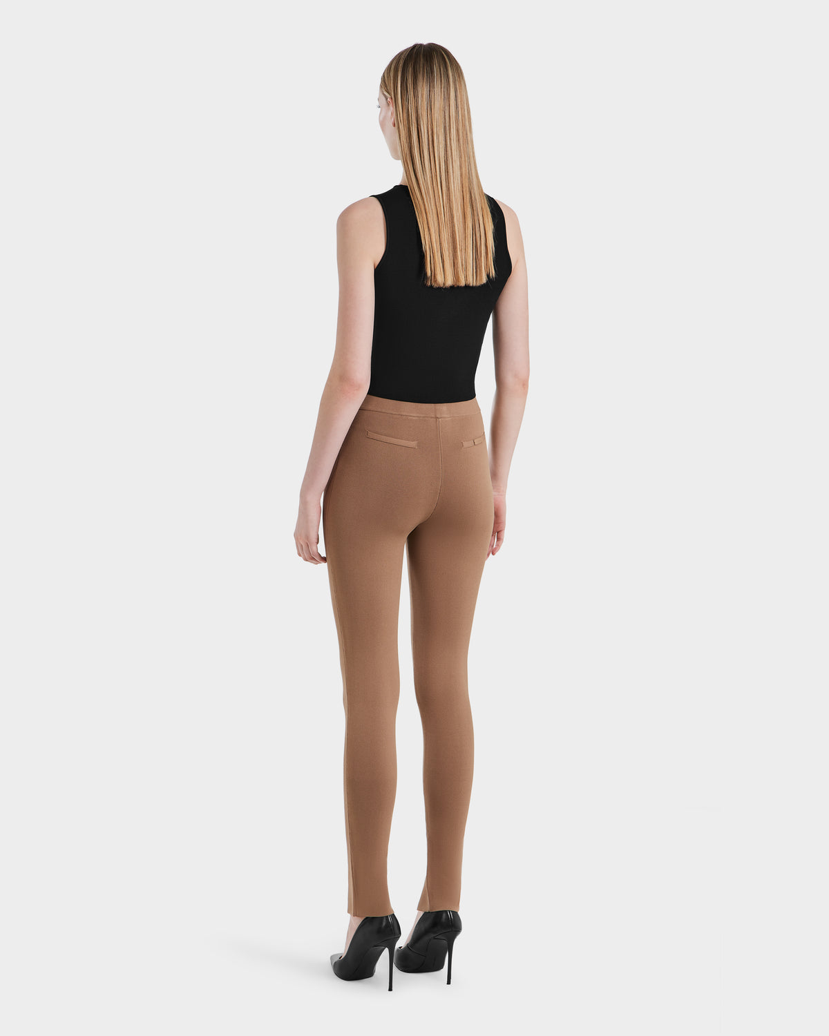 A woman with straight blonde hair, in a sleeveless black top, Rudsaks camel MATHILDA - 8524515 ankle slit leggings, and black heels stands facing away against a plain white background.
