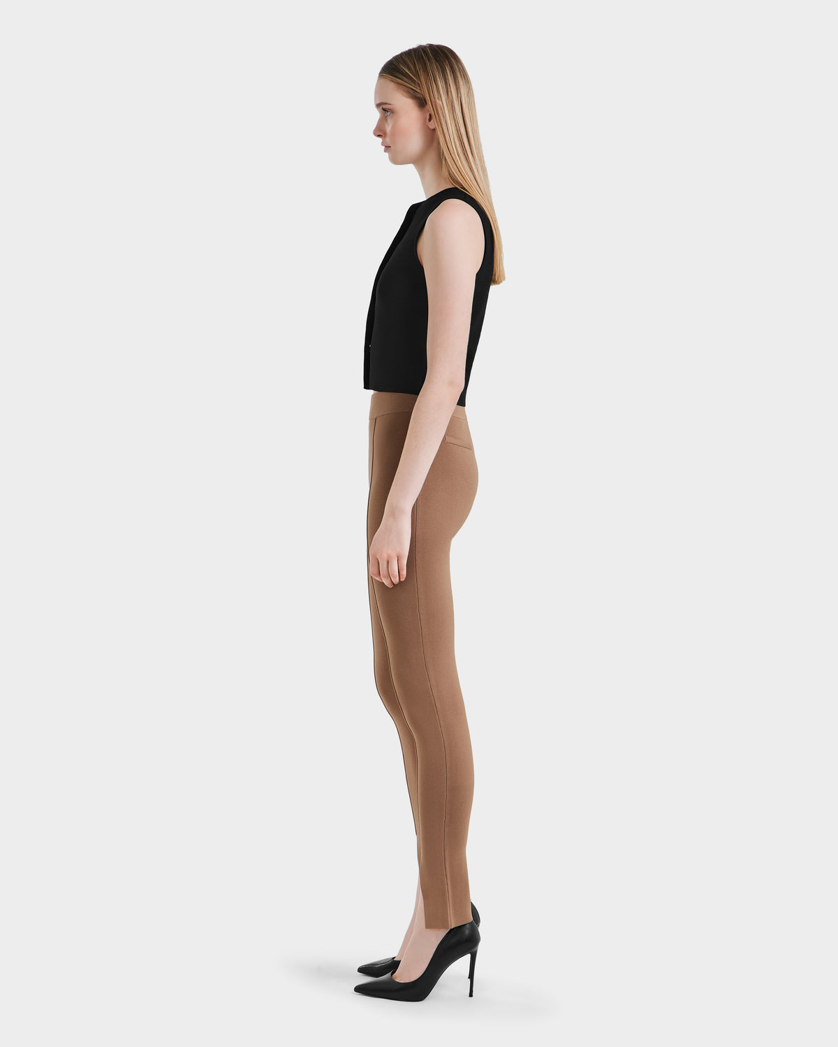 A woman stands in profile against a plain white background, wearing a sleeveless black top, Rudsak MATHILDA - 8524515 Camel ankle slit leggings, and black high-heeled shoes.