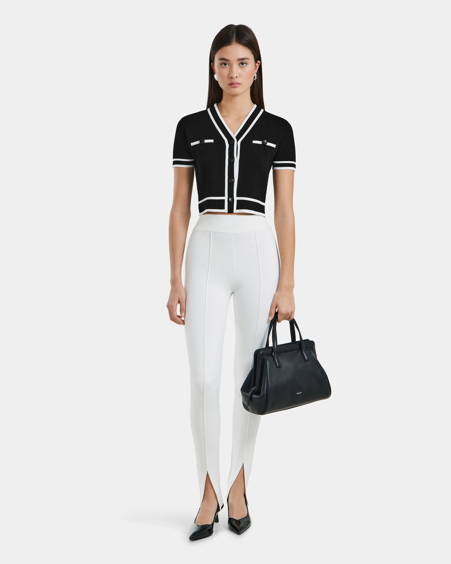 A woman stands against a white background wearing short-sleeved black cropped cardigan, Rudsak MATHILDA - 8524515 Iceland ankle slit leggings, black pointed heels, and holding a black handbag.