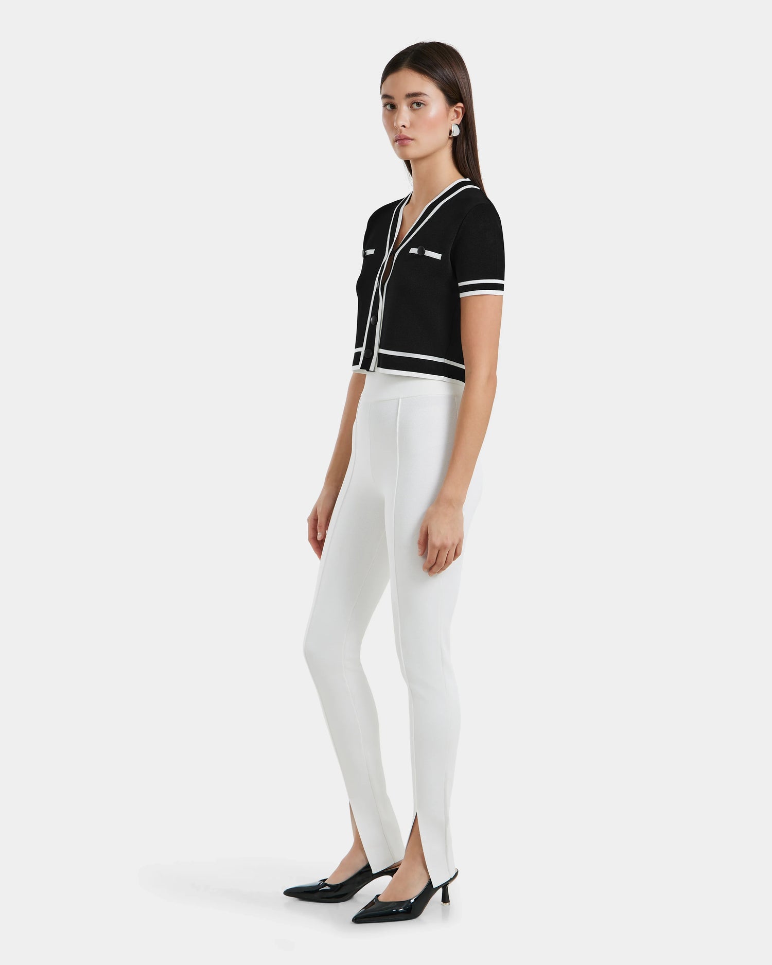 A woman poses against a plain background in the MATHILDA - 8524515 Iceland black short-sleeve top by Rudsak, featuring white trim and buttons, paired with high-waisted front ankle-slit leggings and black pointed-toe heels.