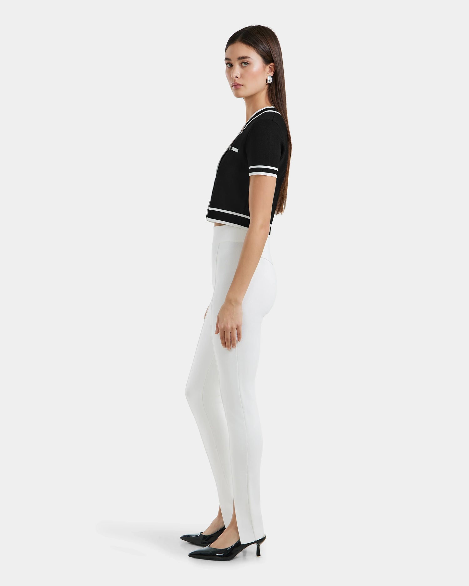 A woman stands sideways against a plain white background, wearing black short-sleeve top with white trim, MATHILDA - 8524515 Iceland leggings by Rudsak featuring front ankle slits, and black low-heeled shoes.