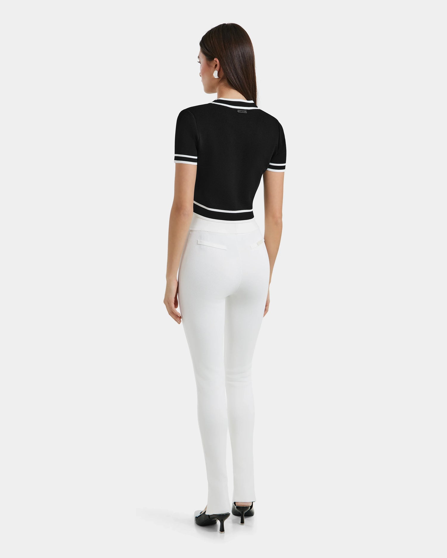 A woman with straight brown hair wears the Rudsak MATHILDA - 8524515 Iceland black short-sleeve top with white trim and fitted white knit leggings, styled with black heels against a plain white background.