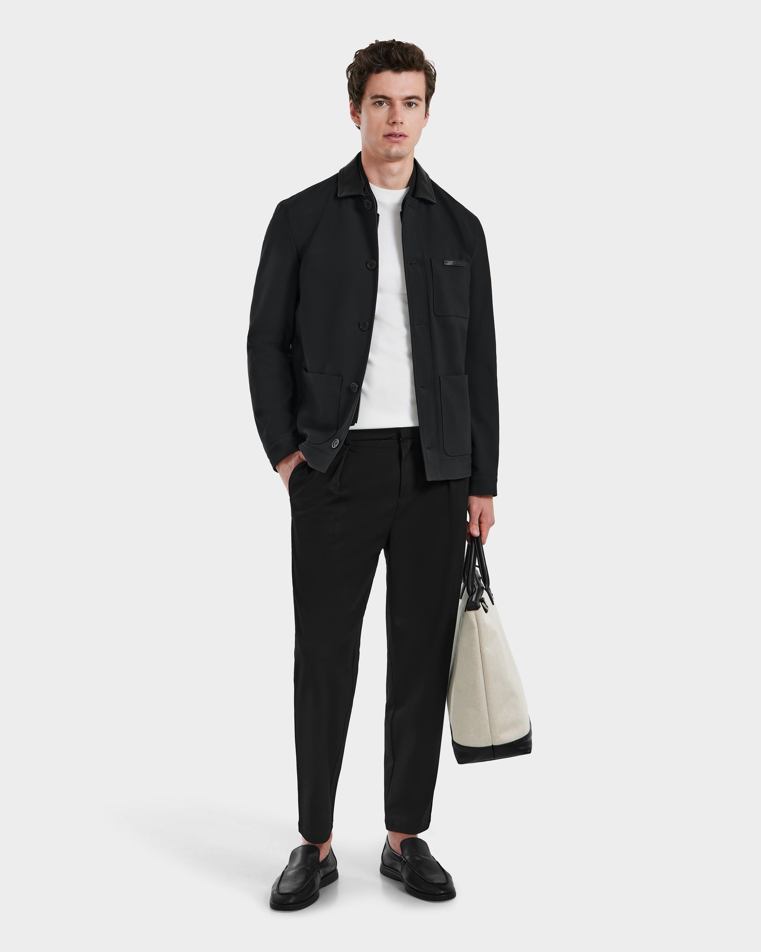 A young man stands against a plain background wearing Rudsaks MATTHEW - 6125004 Black blazer, a white shirt, black pants, and black loafers, holding a large white and black tote bag in his left hand.