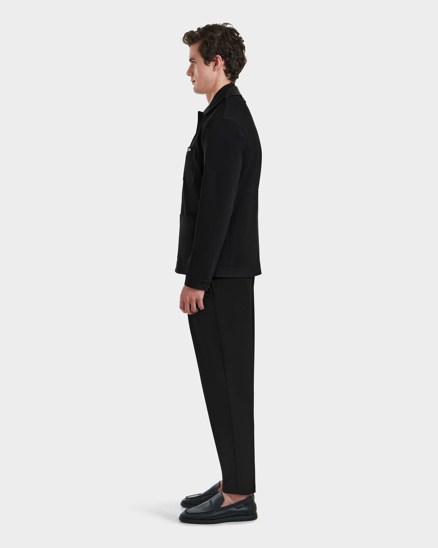 A man stands in profile against a white background, wearing the Rudsak MATTHEW - 6125004 Black blazer, paired with black pants and shoes. His arms are relaxed at his sides as he faces to the left.