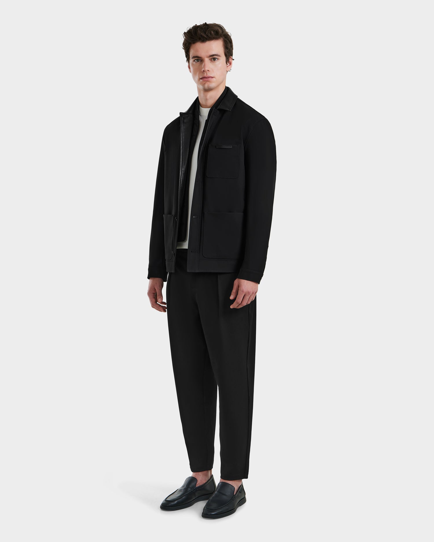 A man stands against a white background wearing the Rudsak MATTHEW - 6125004 Black tailored blazer with technical fabric, a white shirt, black trousers, and black loafers, looking directly at the camera with a neutral expression.