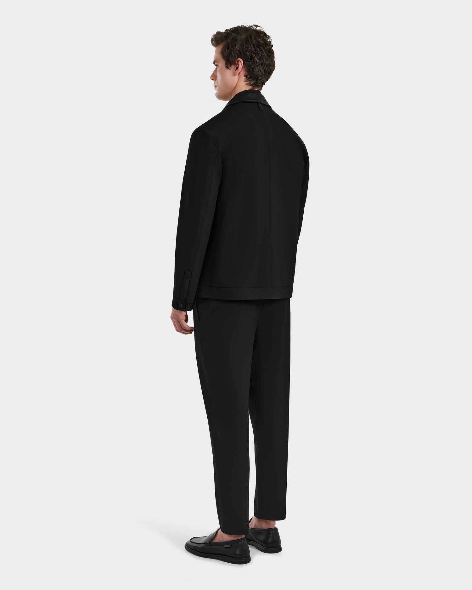 A man stands facing away from the camera, wearing Rudsak’s MATTHEW - 6125004 Black tailored blazer and trousers, paired with black loafers against a plain light gray background.