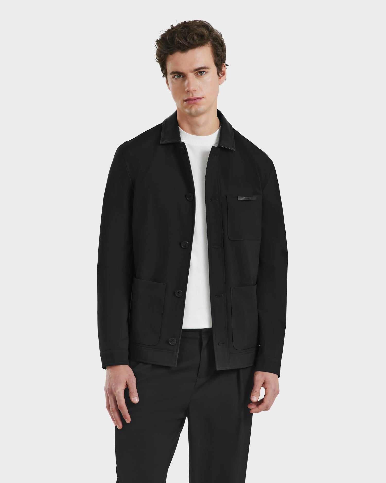 A young man with short brown hair models the Rudsak MATTHEW - 6125004 Black tailored blazer-style shirt jacket over a white shirt and black pants, standing against a plain light gray background.