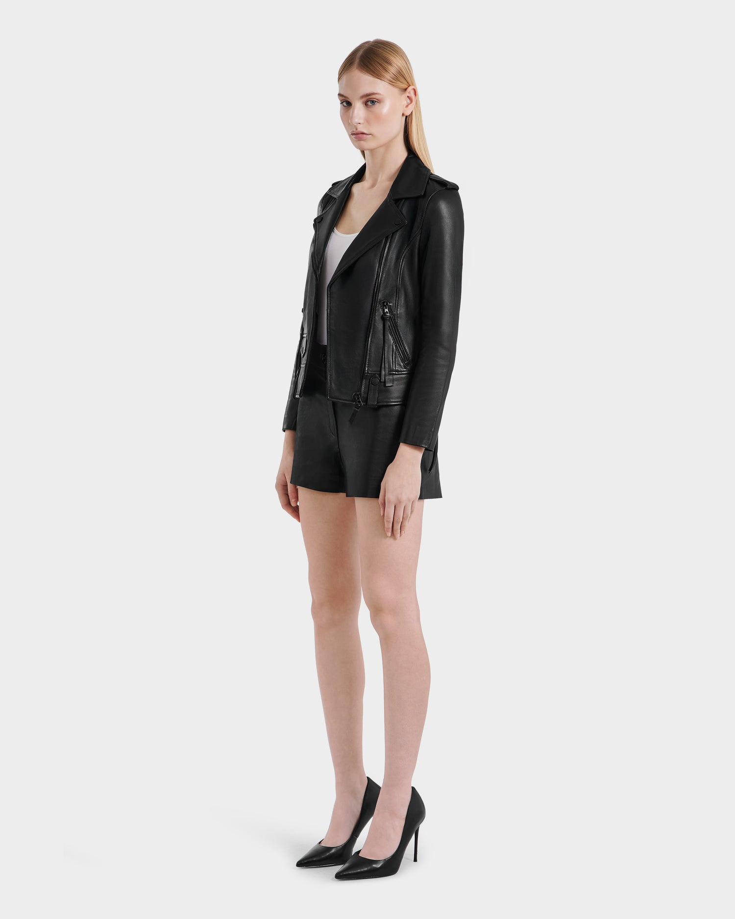 A woman with straight, light brown hair and a neutral expression stands against a white background wearing the Rudsak MERGO - 8122009 BLACK lambskin leather biker jacket, black shorts, a white top, and black high heels.