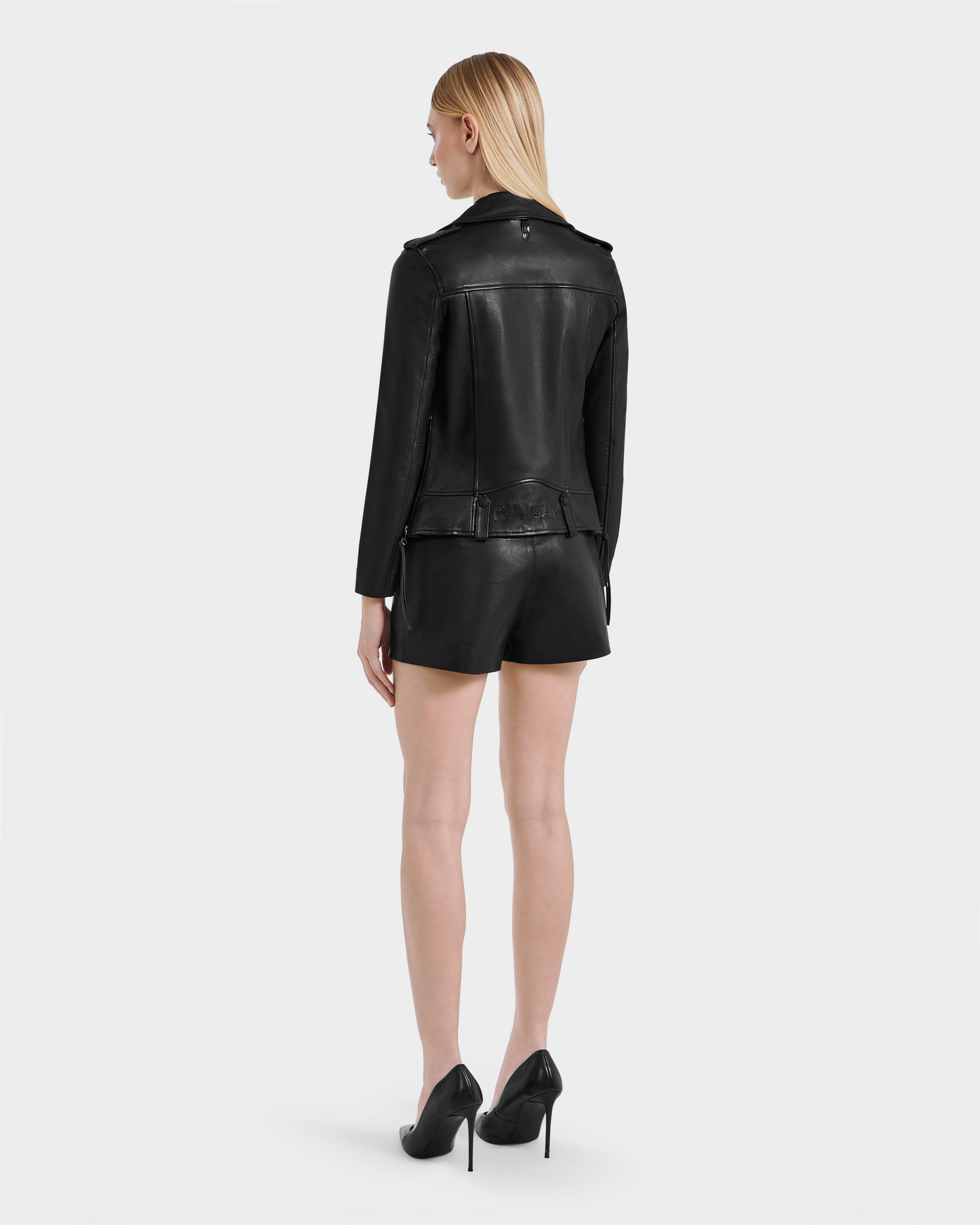 A woman with straight blonde hair stands facing away, wearing the Rudsak MERGO - 8122009 BLACK sleek black lambskin leather jacket, black leather shorts, and high heels against a plain white background.