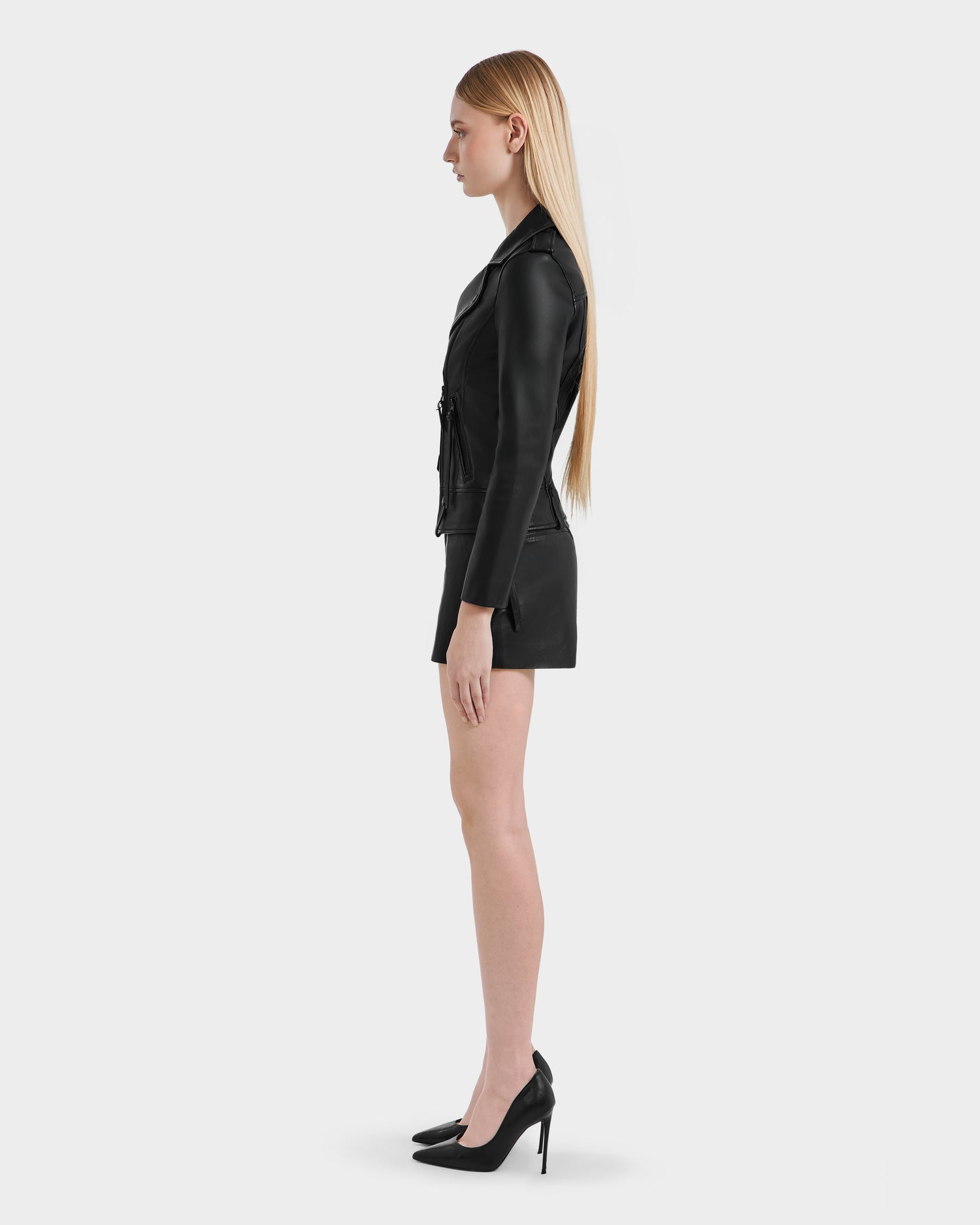 A woman with long, straight blonde hair stands in profile against a plain white background, wearing the Rudsak MERGO - 8122009 BLACK lambskin leather biker jacket, paired with a black mini skirt and high heels.
