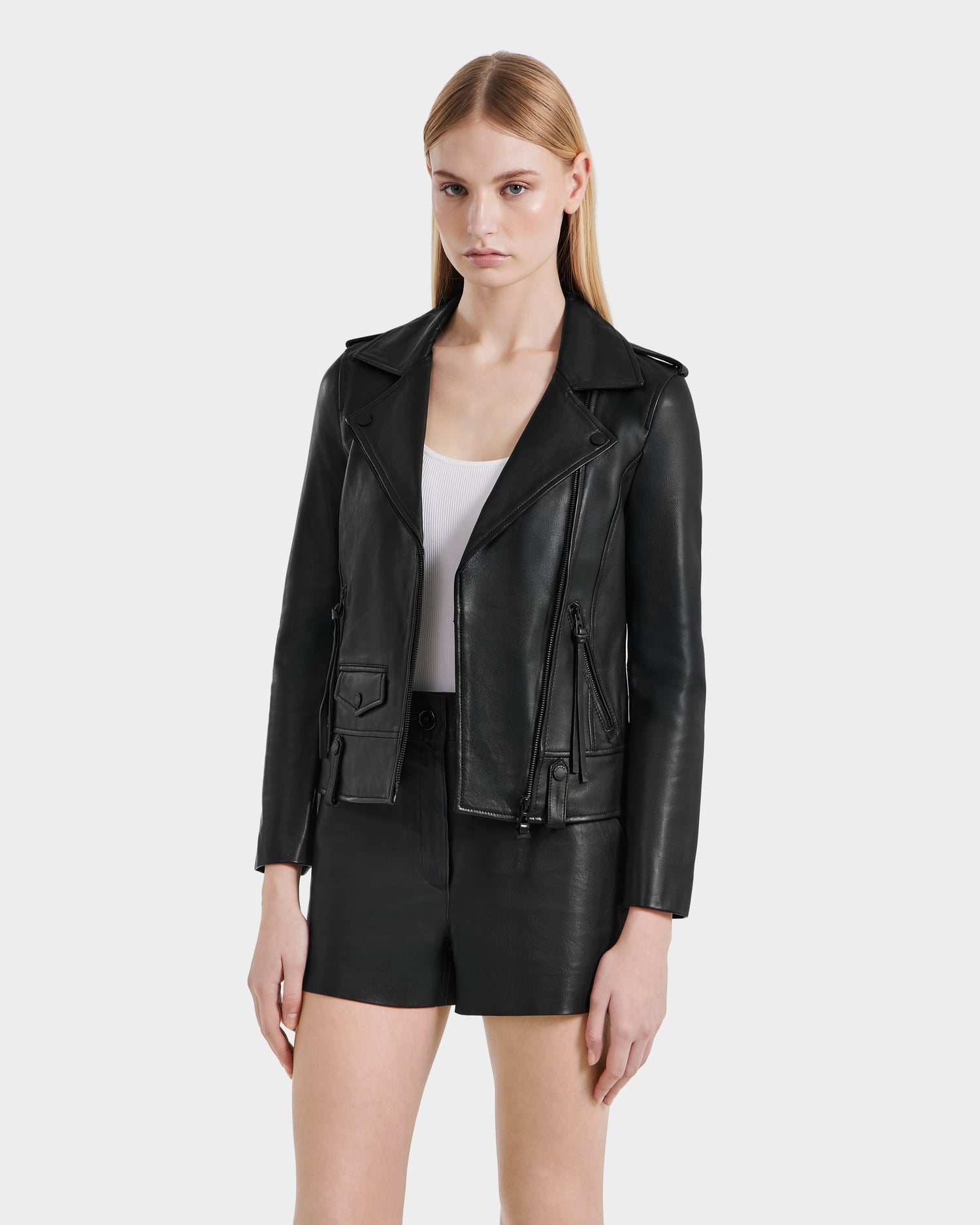 A woman with straight blonde hair wears the Rudsak MERGO - 8122009 BLACK lambskin leather biker jacket over a white top and black shorts, standing against a plain light background.