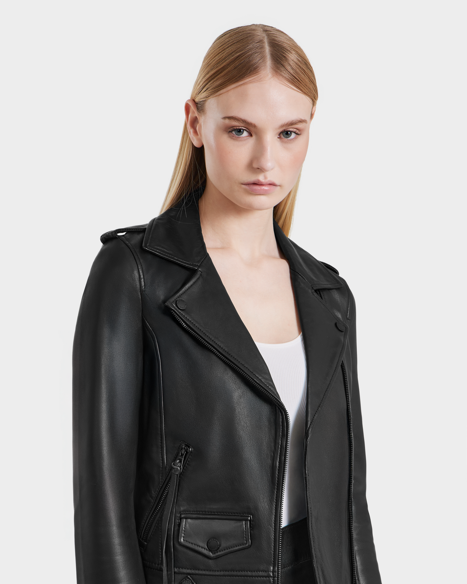 A woman with straight blonde hair wears the Rudsak MERGO - 8122009 BLACK lambskin biker jacket over a white top, standing against a light gray background and looking directly at the camera with a neutral expression.
