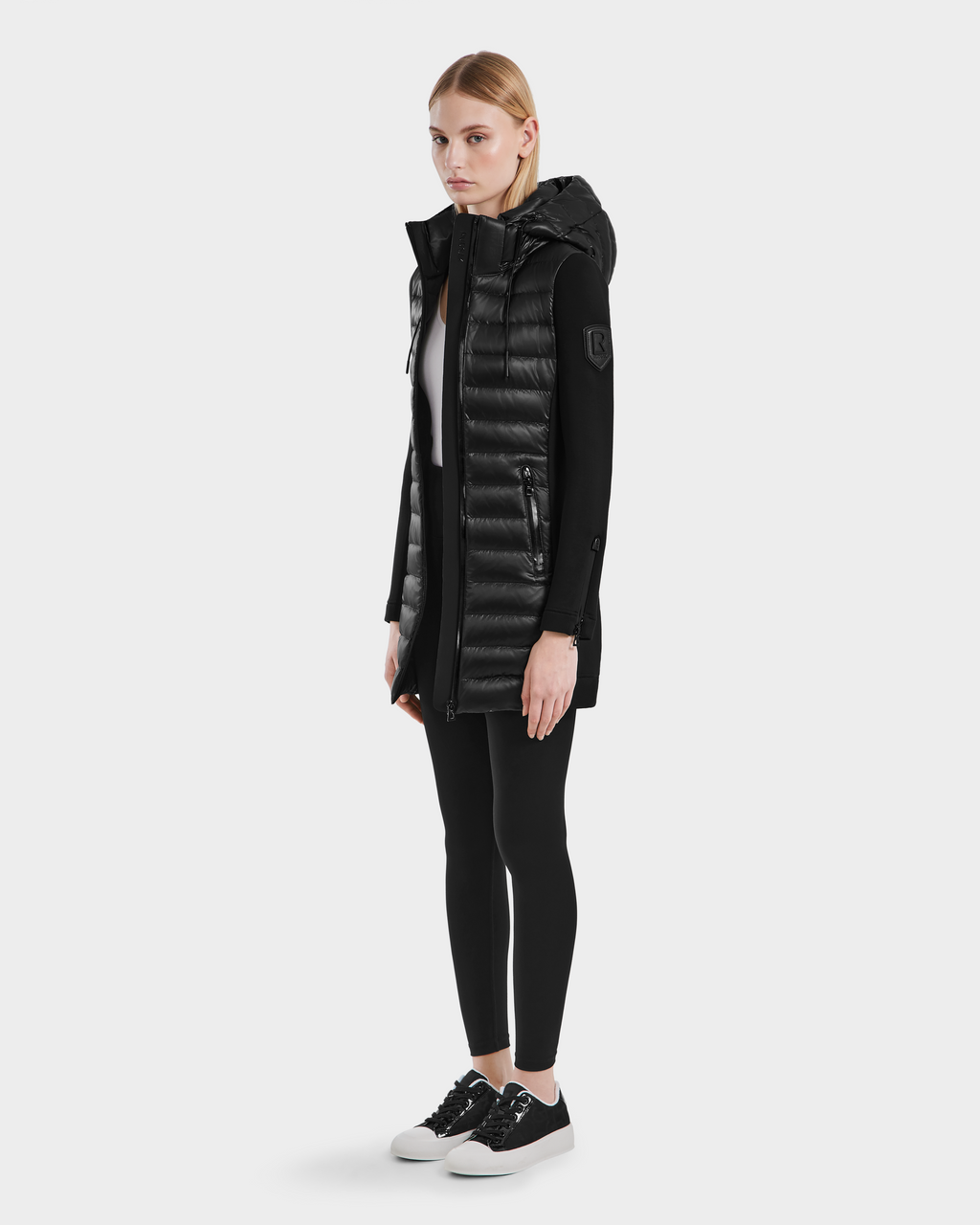 A woman faces forward wearing the Rudsak MERINA - 8121006 BLACK lightweight hooded puffer vest over a black long-sleeve top, black leggings, and black-and-white sneakers against a plain white background.
