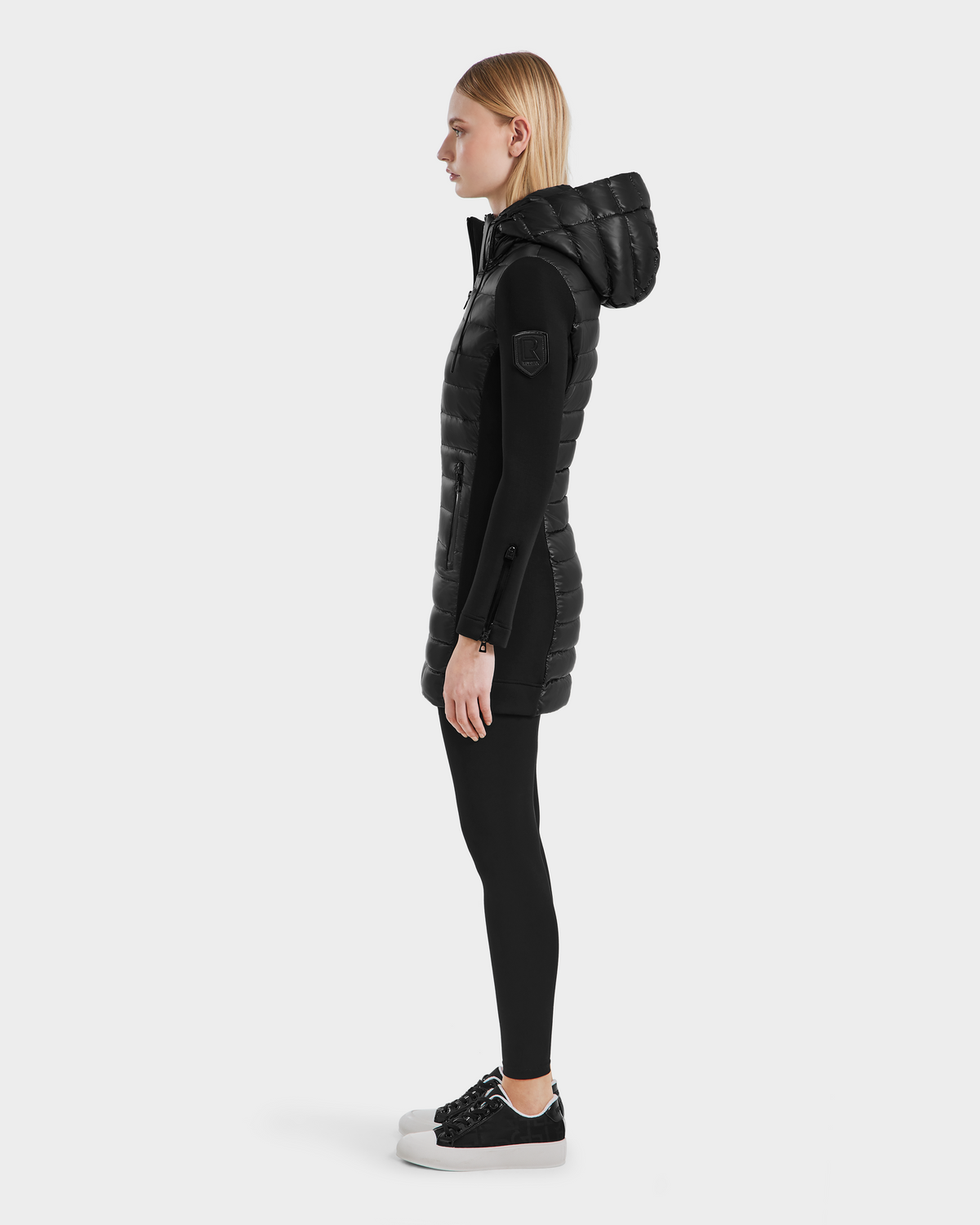 A woman in profile wears the Rudsak MERINA - 8121006 BLACK hooded puffer vest over a black long-sleeve top, leggings, and black-and-white sneakers, standing against a plain white background.