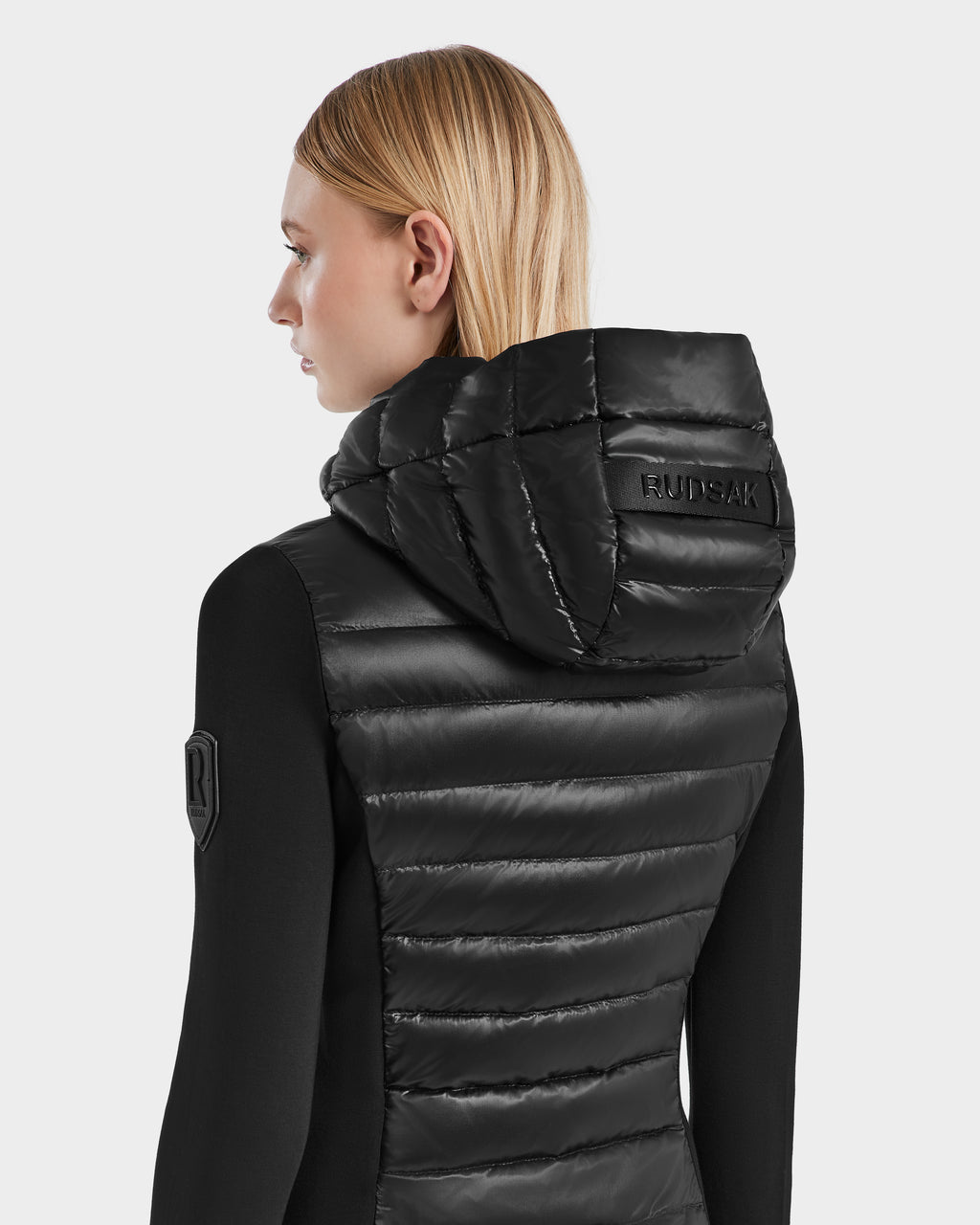 A woman with straight blonde hair is seen from the back wearing the Rudsak MERINA - 8121006 BLACK womens light puffer vest with RUDSAK embossed on the hood, paired with a black long-sleeve top featuring an arm patch.