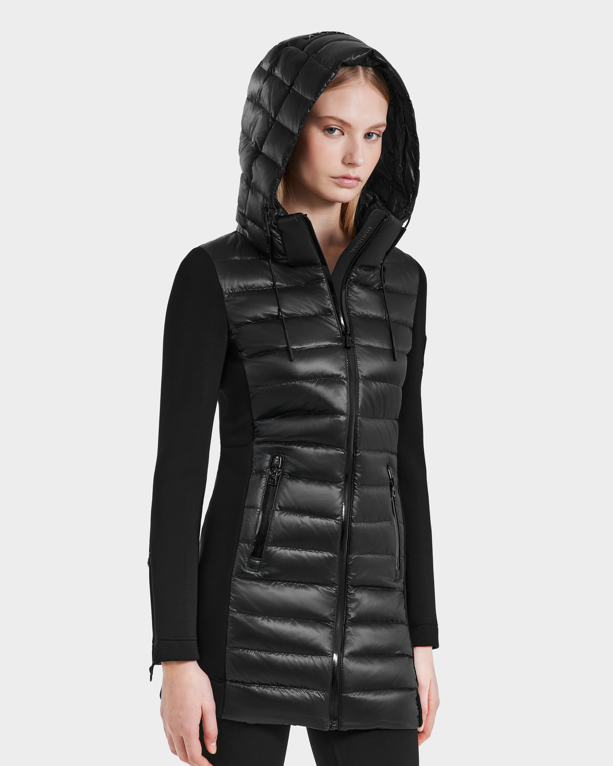 A woman wears the Rudsak MERINA - 8121006 BLACK quilted down jacket with zippered pockets and long sleeves, standing against a plain light gray background.
