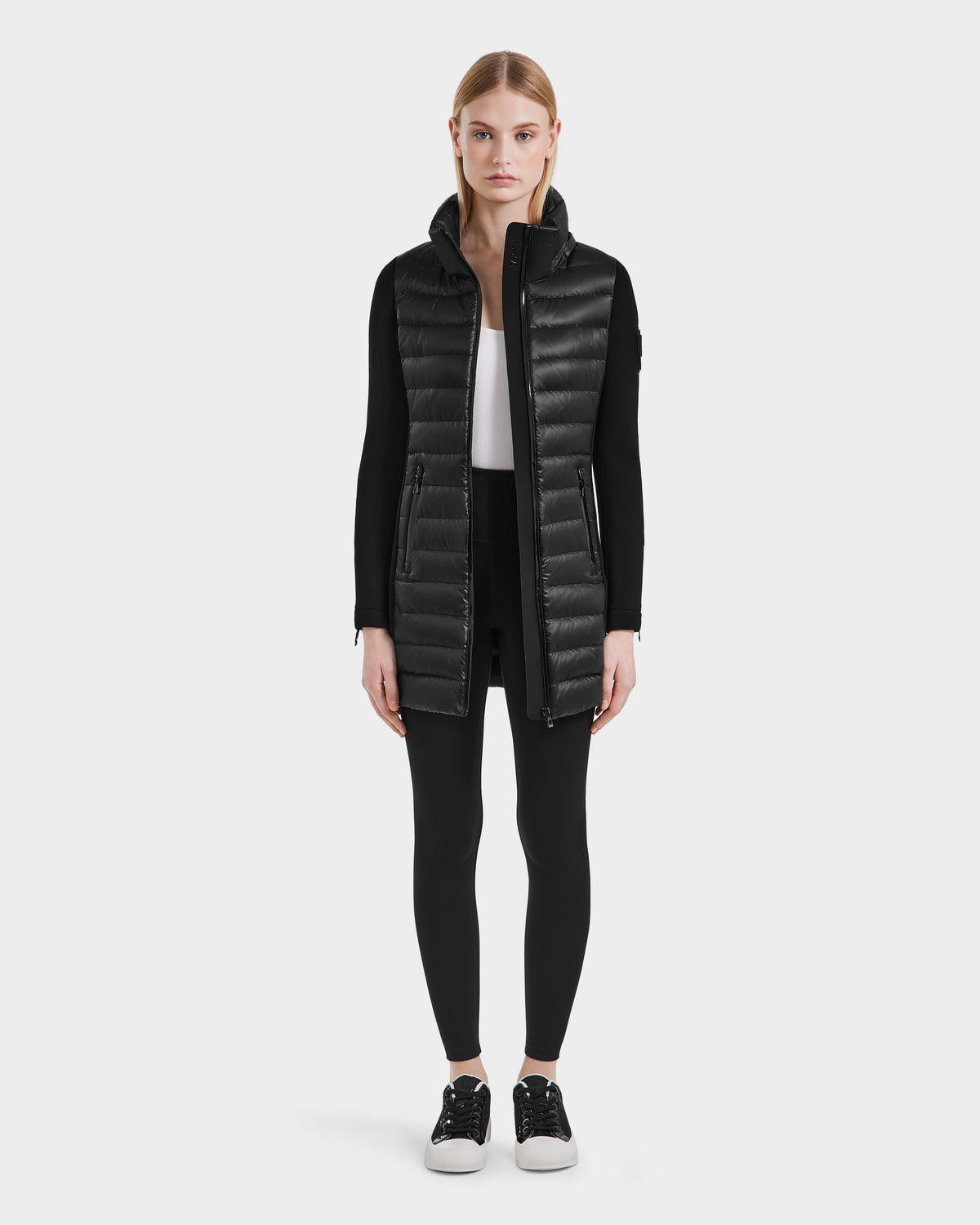 A woman stands facing forward in the Rudsak MERINA - 8121006 BLACK lightweight jacket over a white top, black leggings, and black sneakers, set against a plain light background.