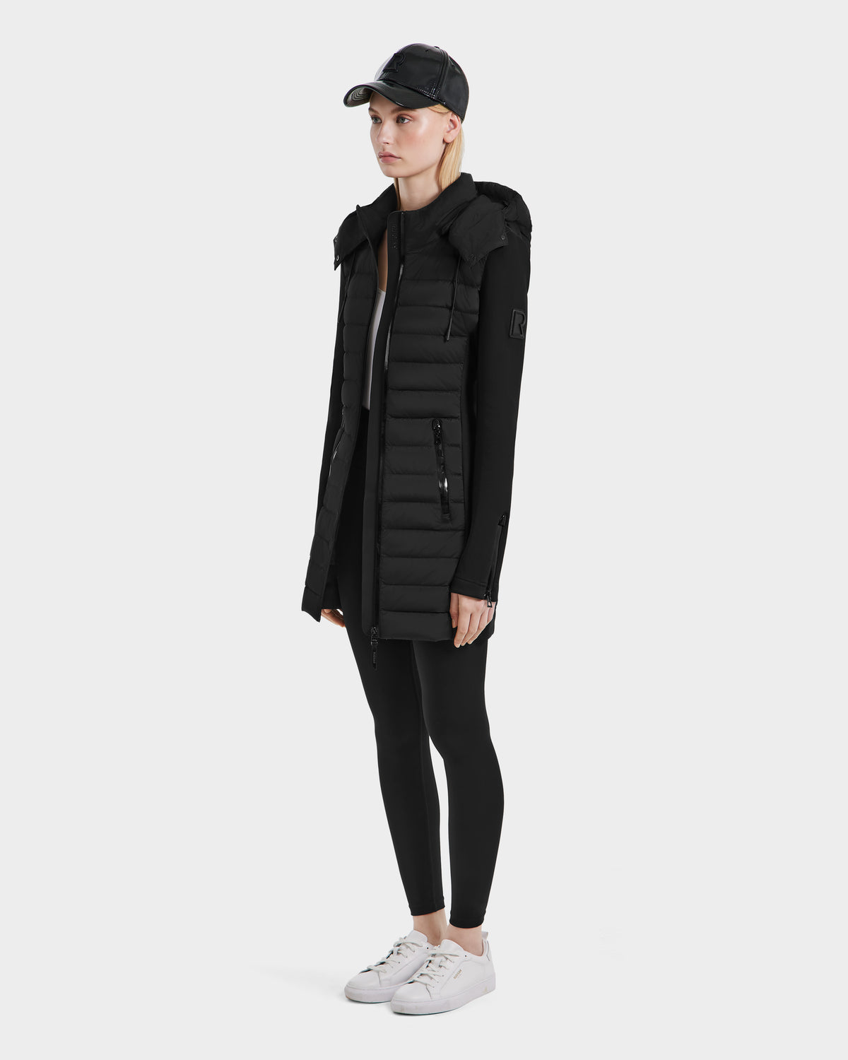 A person stands against a plain white background wearing the Rudsak MERINA - 8124826 Black womens light quilted jacket, paired with black leggings, a black baseball cap, and white sneakers.