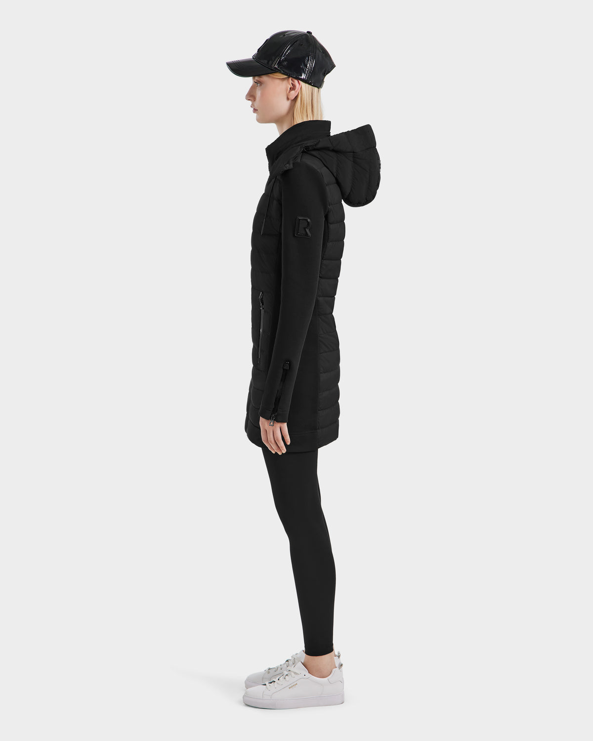 A person stands in profile wearing the Rudsak MERINA - 8124826 Black quilted down jacket with a hood, paired with black leggings, a black baseball cap, and white sneakers against a plain light background.