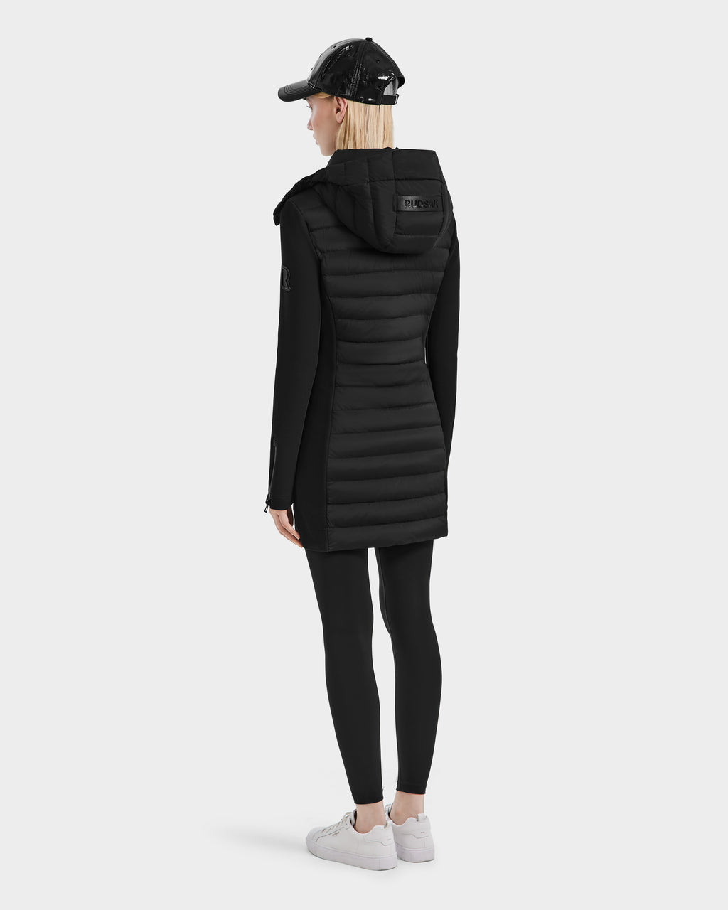 A woman faces away, wearing the Rudsak MERINA - 8124826 Black quilted down jacket with black leggings, a black cap, and white sneakers against a plain white background.