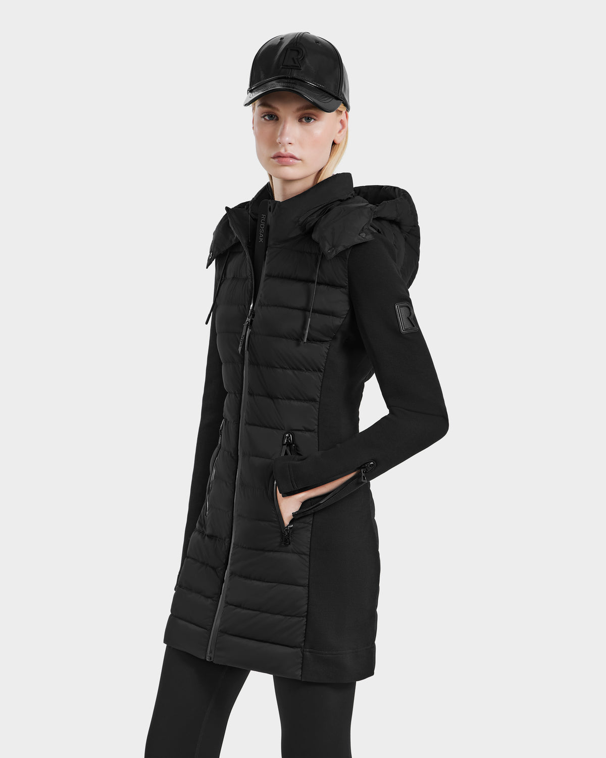 A person wearing the Rudsak MERINA - 8124826 Black quilted down jacket, paired with black leggings and a black baseball cap, stands with hands in pockets against a plain light gray background.
