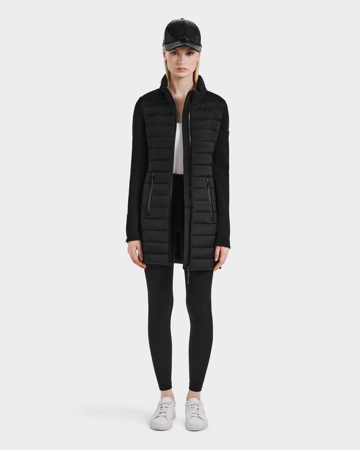 A woman faces forward against a plain light background, wearing the Rudsak MERINA - 8124826 Black long quilted down jacket, paired with a black cap, black leggings, white top, and white sneakers.