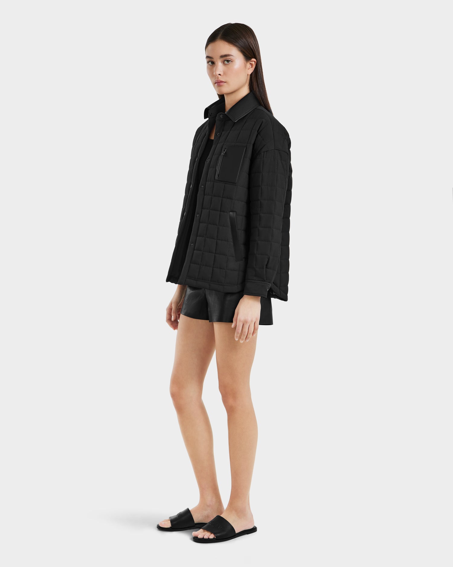 A woman with straight brown hair and a neutral expression stands against a plain background, wearing the Rudsak NATALYA - 8125030 Black vegan leather jacket paired with black shorts and slide sandals. Her arms rest relaxed at her sides.