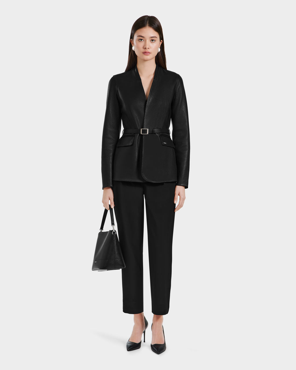 A woman showcases minimalist sophistication in the Rudsak PAMILA - 8125051 Black leather blazer with a black belt, black pants, pointed-toe heels, and a black handbag; her sleek dark hair contrasts the plain light background.